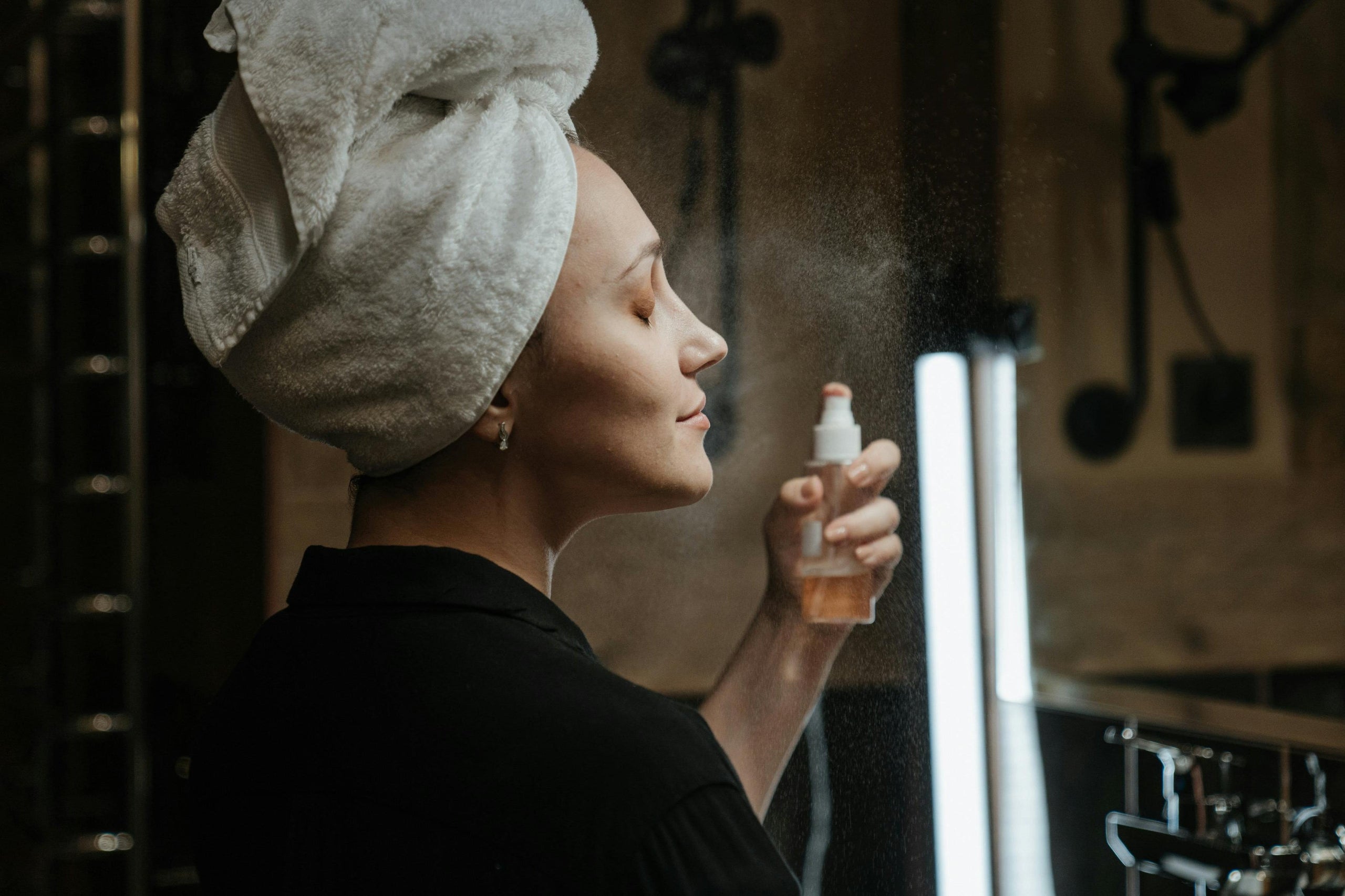 Woman with a towel on her head holding a hair product spray bottle in a bathroom setting.slider_item_zRzGTJ